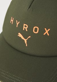 Cappello verde oliva con tessuto testurizzato, con la scritta "HYROX" in arancione e un piccolo logo Puma arancione sul davanti.
