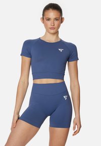 Young woman wearing matching blue crop top and high-waisted shorts with white logo, standing against plain white background.