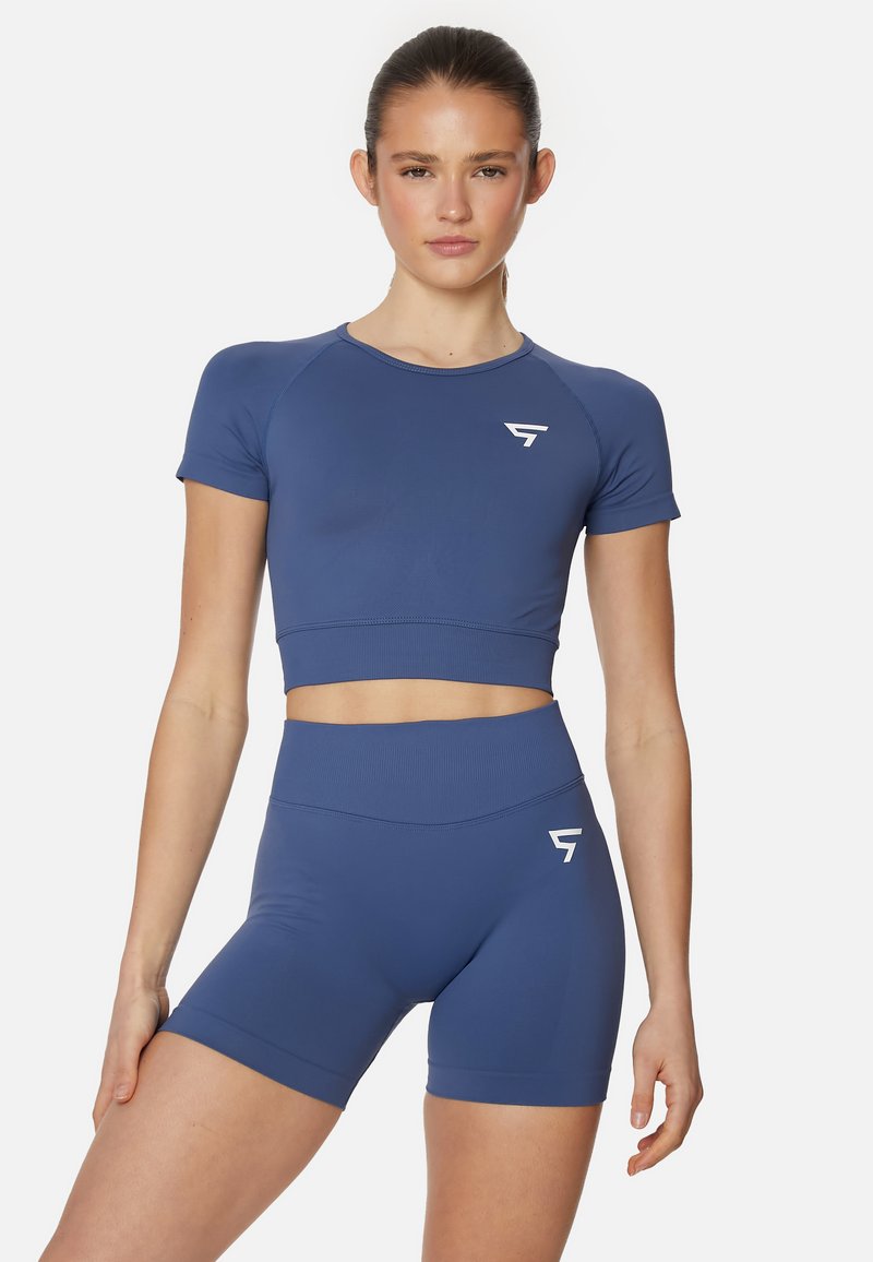 Mladá žena v modrom crop top a vysokých šortkách s bielym logom, stojaca na pozadí z jednofarebnej bielej.