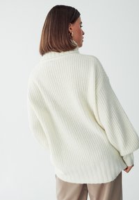 Maglione bianco a maglia con texture a coste e collo alto, caratterizzato da spalle scese e una vestibilità rilassata, abbinato a pantaloni beige chiaro.