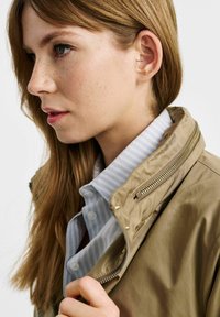 Jasno brązowa kurtka z zamkiem błyskawicznym przy kołnierzu, noszona na niebieskiej i białej koszuli w paski zapinanej na guziki. Gładki materiał i minimalne detale sprzętowe.