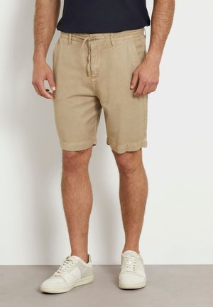 Beżowe lniane szorty z elastycznym pasem i sznurkiem do regulacji. Posiadają podwinięte mankiety i kieszenie. Połączone z białymi trampkami.
