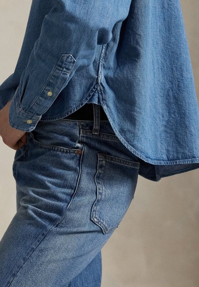 Persoon draagt blauwe spijkerbroek en een blauw denim overhemd dat niet is ingestopt, staand tegen een effen lichte achtergrond.