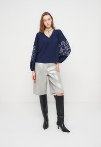 Γυναίκα που φοράει μπλε σκούρο μπλούζα με κεντήματα λουλουδιών στα μπαλόν μανίκια, ασημένια σορτς μήκους μέχρι το γόνατο και μαύρες μπότες με τακούνι μέχρι το γόνατο.