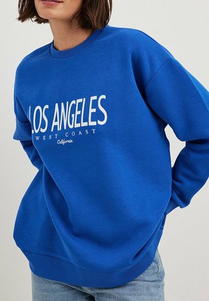 Femme portant un sweat-shirt bleu avec le texte « LOS ANGELES WEST COAST California », associé à un jean bleu clair sur un fond uni.