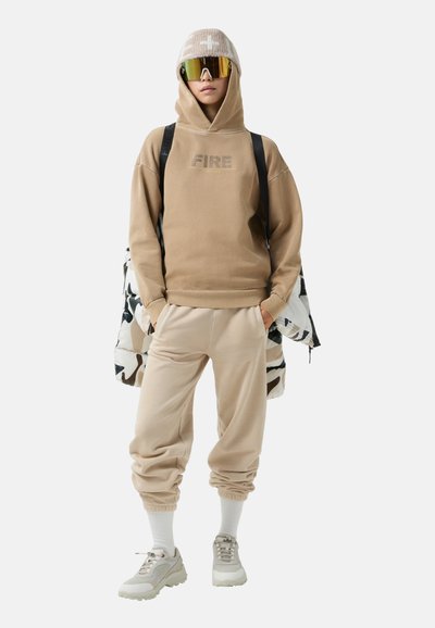 Beżowa bluza z kapturem z grafiką "FIRE", zestawiona z pasującymi joggerami, białymi skarpetkami i masywnymi sneakersami. Model trzyma wzorzystą kurtkę i nosi okulary przeciwsłoneczne z lustrzanymi soczewkami.