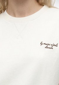 Gros plan d'un sweat-shirt blanc à col rond avec un petit texte cursif marron sur la poitrine indiquant "by marc o'polo denim".
