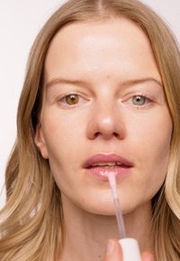 Applicatore di lucidalabbra trasparente tenuto vicino alle labbra, con una consistenza liscia e rosa. Il modello ha due occhi di colore diverso: uno marrone e uno verde.