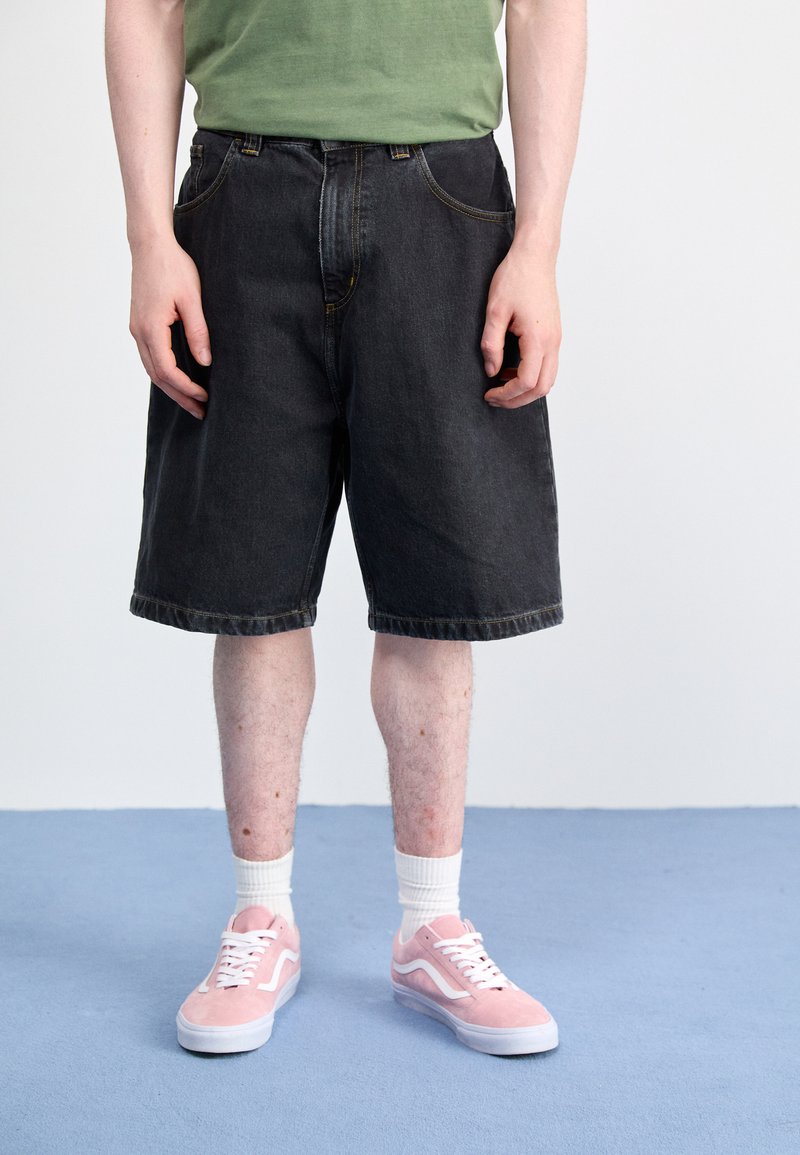 Shorts de mezclilla negra con un corte holgado, que presentan costuras amarillas y bolsillos. Combinados con zapatillas de ante rosa y calcetines blancos.