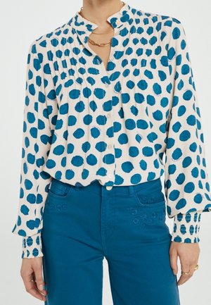 Biała bluzka w niebieskie groszki, plisowany fason i zapinany na guziki dekolt. W połączeniu z turkusowymi spodniami ozdobionymi haftowanymi kwiatowymi detalami.