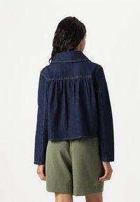 Veste en denim courte de couleur bleu foncé avec un design plissé à l'arrière, col, et surpiqûres contrastées. Associée à un short vert olive.