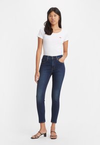 Hoch geschnittene dunkelblaue Jeans mit einer taillierten Form, ergänzt durch ein schlichtes weißes T-Shirt mit einem kleinen roten Logo. Schwarze Riemchensandalen.