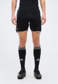 Άτομο που φοράει μαύρο αθλητικό σορτς, μαύρες κάλτσες Adidas μέχρι το γόνατο με λευκές ρίγες και μαύρα ποδοσφαιρικά παπούτσια, στέκεται σε λευκό φόντο.