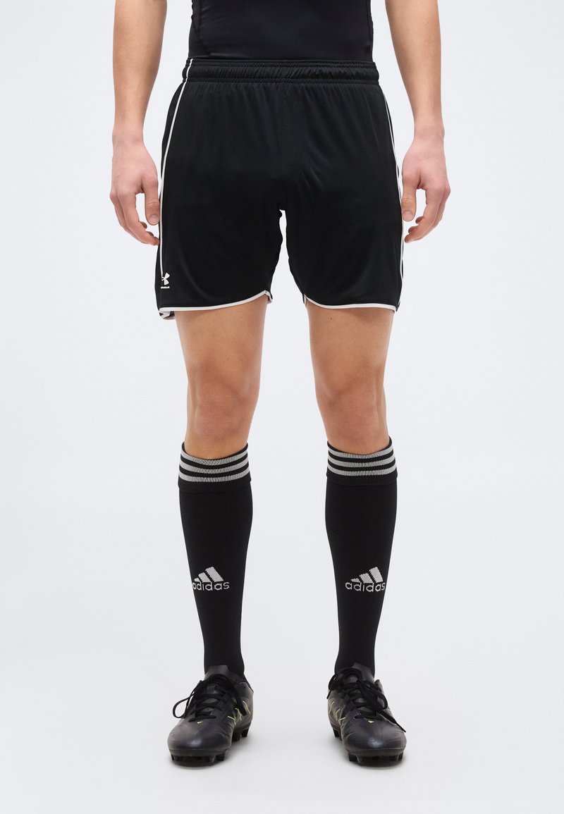 Άτομο που φοράει μαύρο αθλητικό σορτς, μαύρες κάλτσες Adidas μέχρι το γόνατο με λευκές ρίγες και μαύρα ποδοσφαιρικά παπούτσια, στέκεται σε λευκό φόντο.