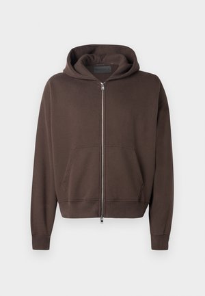 Smeđa zip-up hoodie napravljena od mekog materijala, s velikom kapuljačom, prednjim kengur-pocketom i rebrastim manžetama i rubom.