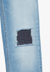 Los jeans de mezclilla azul claro presentan un parche desgastado con bordes deshilachados, revelando mezclilla azul oscura debajo. Diseño clásico de cinco bolsillos.