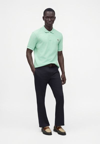 Homme debout portant un polo vert menthe à manches courtes, pantalon foncé, ceinture marron et chaussures habillées bicolores marron et beige.