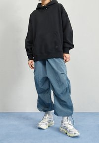 Sudadera negra extragrande con bolsillo frontal, combinada con pantalones cargo texturizados azul claro y zapatillas plateadas con detalles en blanco.