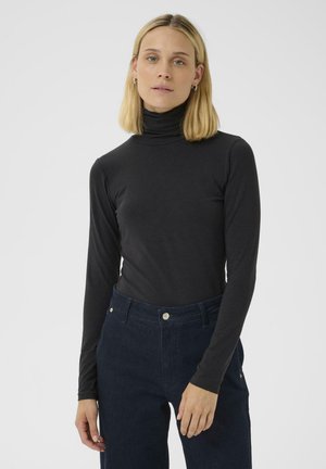 Chandail à col roulé noir à manches longues en tissu lisse, associé à un pantalon taille haute bleu foncé avec poches avant et détails en métal argenté.