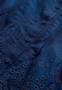 Mörkblått tyg med broderade blomster mönster och hålmönsterdetaljer. Har matchande blå knappar längs framsidan. Texturerad och lager-på-lager design.