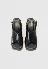 Sorte åpen-tå sandaler med kryssstropper, glatt lærtekstur og en polstret såle. Har glidelås på siden.
