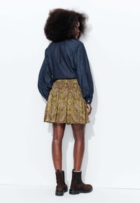 Chemise en denim à manches longues associée à une jupe jaune et marron à motifs, portée avec des bottines brunes. Vue de dos sur fond blanc.