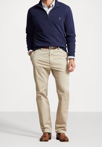 Suéter azul marino con cuello con cremallera, chinos beige, zapatos de cuero marrón y una camisa a cuadros en azul claro debajo. Logo de Polo en el suéter.