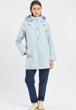 Femme portant un imperméable à capuche bleu clair, un pantalon bleu marine et des chaussures beiges à lacets, debout avec une main dans la poche sur un fond blanc.
