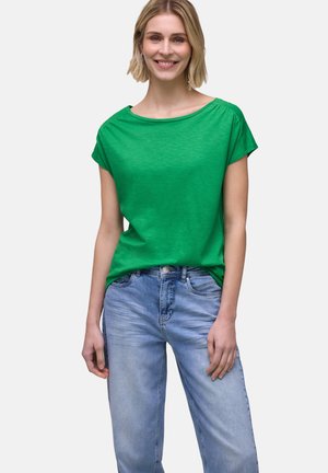 Frau trägt ein grünes Kurzarmshirt und hellblaue Jeans, steht lächelnd vor einem einfarbigen grauen Hintergrund.