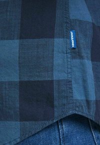 Blå och mörkblå rutigt tygskjorta med synlig sidsöm och "JACK & JONES"-etikett, bärs över blå denimjeans.