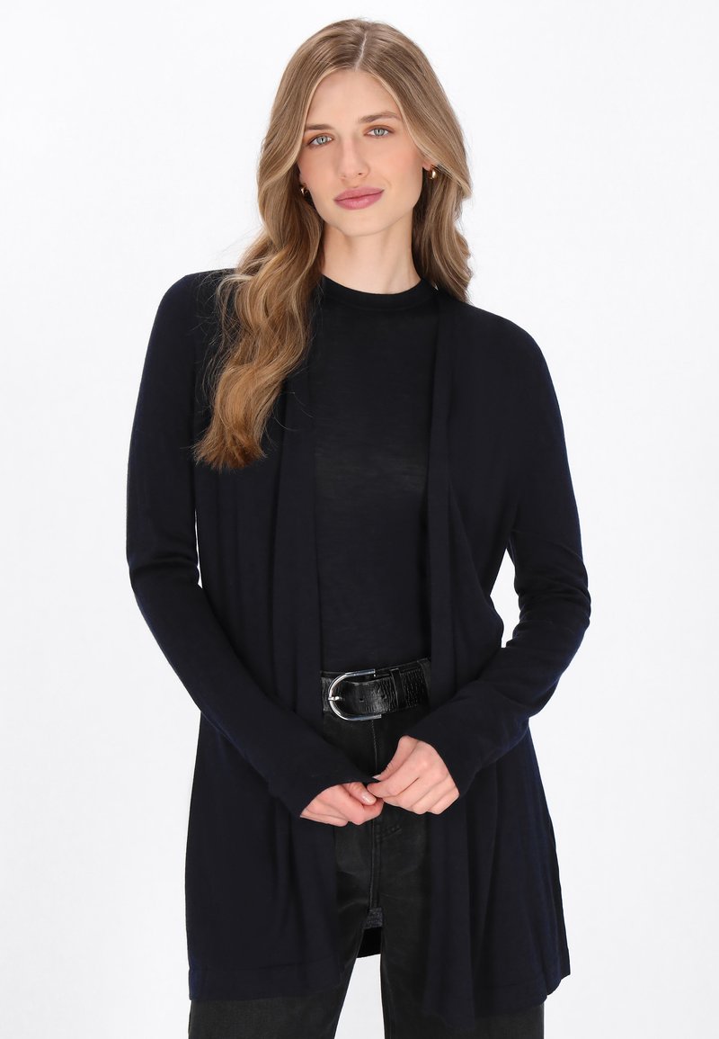 Marineblaues langes Cardigan mit offenem Front, langen Ärmeln und weichem Material. Über einem dunklen T-Shirt getragen und mit einem schwarzen Gürtel kombiniert.