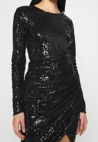 Svart paljettklänning med långa ärmar, texturerad tyg, rynkad design och en sidoöppning, som har en mjuk silhuett och reflekterande detaljer.