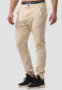 Pantalones cargo beige de corte ajustado, con bolsillos de parche y costuras laterales. La cinturilla lleva el nombre de la marca "INDICODE" en negro.