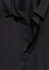 Blusa nera con colletto e pattina bottoni, realizzata in tessuto morbido con una leggera lucentezza. Maniche piegate e vestibilità rilassata.
