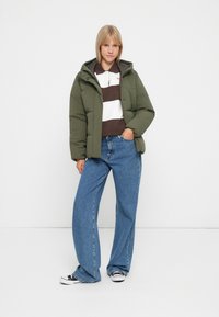 Chaqueta de plumón en verde oliva, con capucha y diseño acolchado. Combinada con un polo de rayas marrón y blanco, pantalones vaqueros de pierna ancha azules y zapatillas.