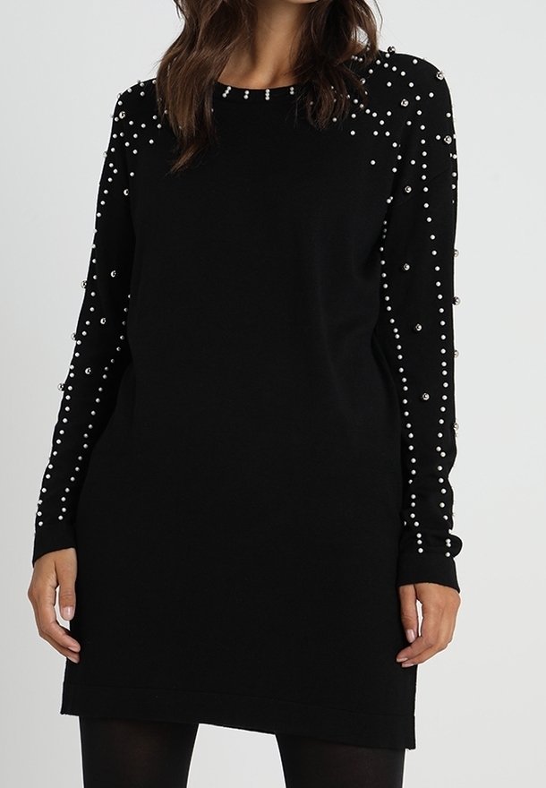 Femme portant une robe noire à manches longues décorée de petites perles blanches sur les manches et les épaules.