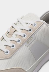 Baskets BOSS blanches et beiges avec panneau latéral texturé, lacets blancs, et bordure en daim autour des œillets, montrant un gros plan de la zone des lacets et du détail du logo.