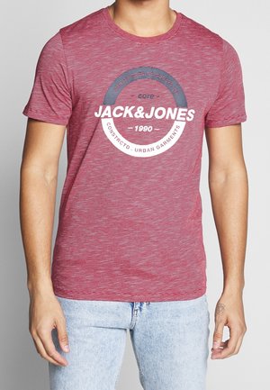 Mann trägt ein rot gestreiftes kurzärmliges T-Shirt mit dem Logo "Jack & Jones 1990", kombiniert mit hellblauen Jeans, steht vor einem grauen Hintergrund.