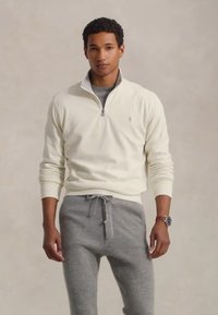 Jonge man in een wit kwart-rits pullover en grijze joggingbroek, staande tegen een effen beige achtergrond.