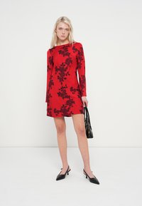 Rdeča obleka z dolgimi rokavi s črnim cvetličnim vzorcem, dolžine do kolen, gladke tkanine in okrogel izrez. Model drži črno torbico.
