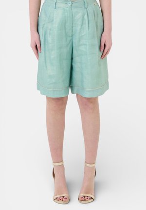 Donna che indossa pantaloncini plissettati a vita alta di colore verde chiaro con orli risvoltati e sandali beige con tacco alto e cinturino alla caviglia, in piedi su sfondo bianco.