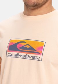 Světle broskvové bavlněné tričko s vícerozměrným vlnovým a horským logem s textem "Quiksilver" v námořnické barvě. Krátké rukávy, kulatý výstřih.