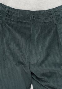 Donkergroene corduroy broek met een gestructureerd, geribbeld oppervlak. Voorzien van een knoopsluiting en een elastische tailleband met geplooide details.