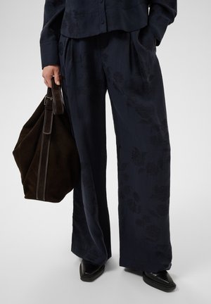 Personne portant un pantalon ample à motifs floraux foncés et des chaussures noires, tenant un grand sac en cuir marron foncé dans une main.