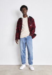 Camicia di flanella rossa e nera con colletto in shearling, t-shirt beige, jeans blu larghi e sneaker bianche. Design semplice, senza motivi visibili.