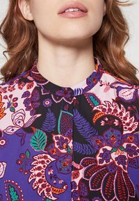 Blusa floreale con una combinazione di colori viola, rosa e nero, con dettagliato motivo e colletto tondo. Tessuto morbido.