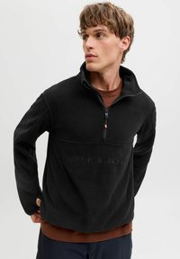 Młody mężczyzna w czarnej polarowej bluzie na zamek z napisem "JACK & JONES", patrzący na bok, z rękami za plecami, na jednolitej jasnej tle.