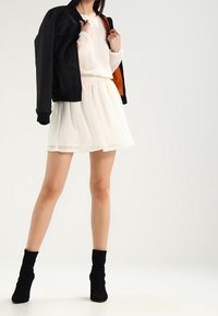 Schwarze Bomberjacke, durchsichtige weiße Langarmbluse, weißer gepunkteter Rock und schwarze Knöchelstiefel. Das Outfit zeichnet sich durch schichtete Texturen und einen taillierten Schnitt aus.