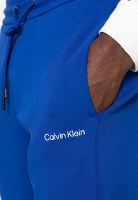 Niebieskie spodnie dresowe z regulowanym troczkiem w pasie, boczną kieszenią oraz białym logo "Calvin Klein" nadrukowanym na lewej udzie. Gładka faktura.