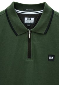 Ciemnozielona koszulka polo z czarnym zamkiem błyskawicznym, czarnym wykończeniem kołnierzyka oraz naszywkami logo "Weekend Offender" na klatce piersiowej i wewnątrz kołnierzyka.